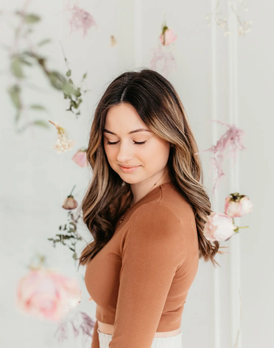A young woman with long, wavy brown hair standing with her eyes closed, smiling softly, surrounded by hanging pink and white flowers against a light background.