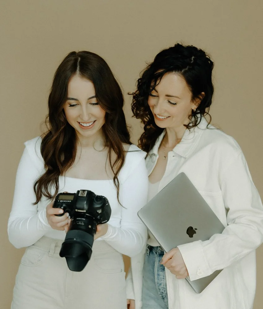 Two women standing close together, smiling, looking at a camera one of them is holding. One woman is holding a camera, the other a laptop.