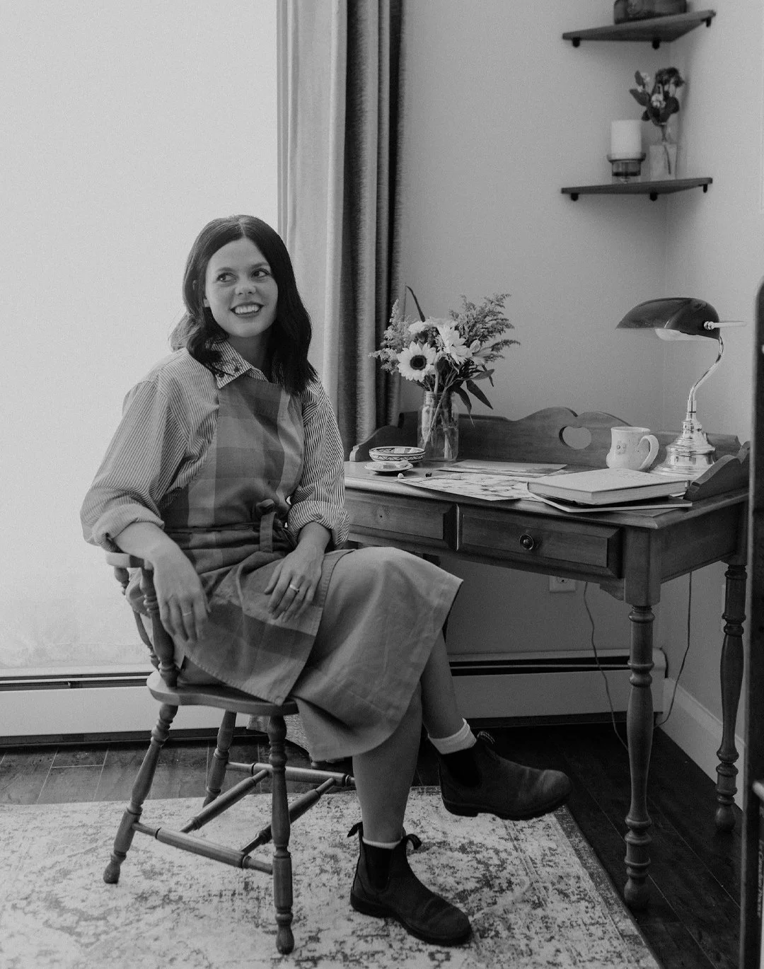 A woman with dark hair sits on a wooden chair in a cozy room, smiling. She is wearing a striped shirt with rolled-up sleeves, an apron, a skirt, and boots. There is a wooden desk with a lamp, a cup, a vase with flowers, and some papers behind her. A window with curtains is on her left and two shelves with decor are on the wall behind her.