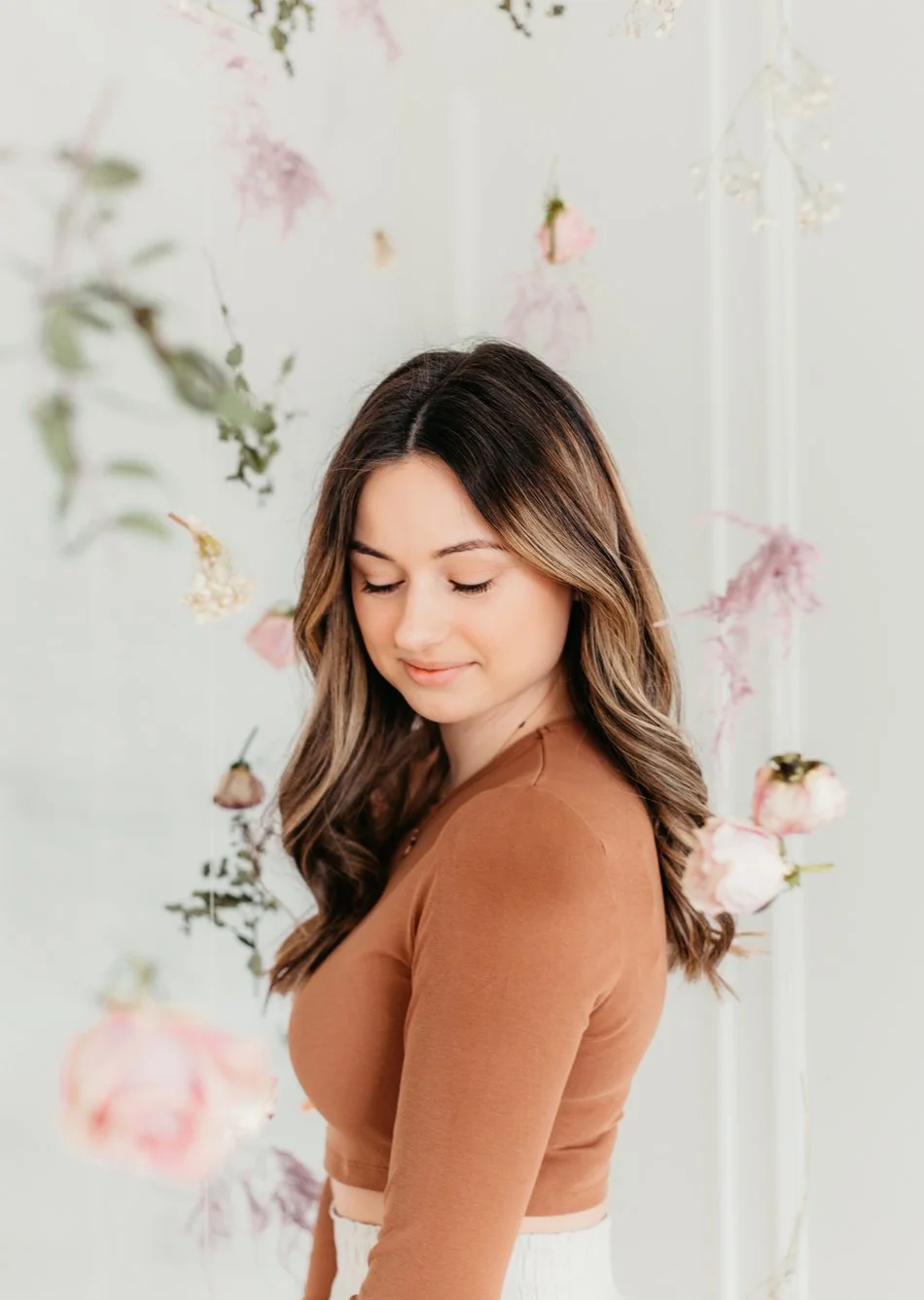 A woman with long brown hair, wearing a brown top, standing amidst hanging pink and white flowers, with her eyes closed and a gentle smile.