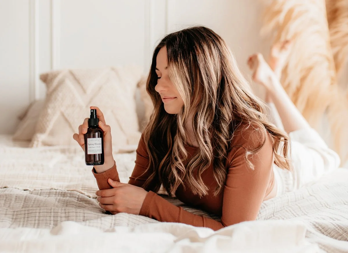 Woman with long wavy hair relaxing on bed, holding a spray bottle labeled 'Spring Blossom' in a cozy bedroom.
