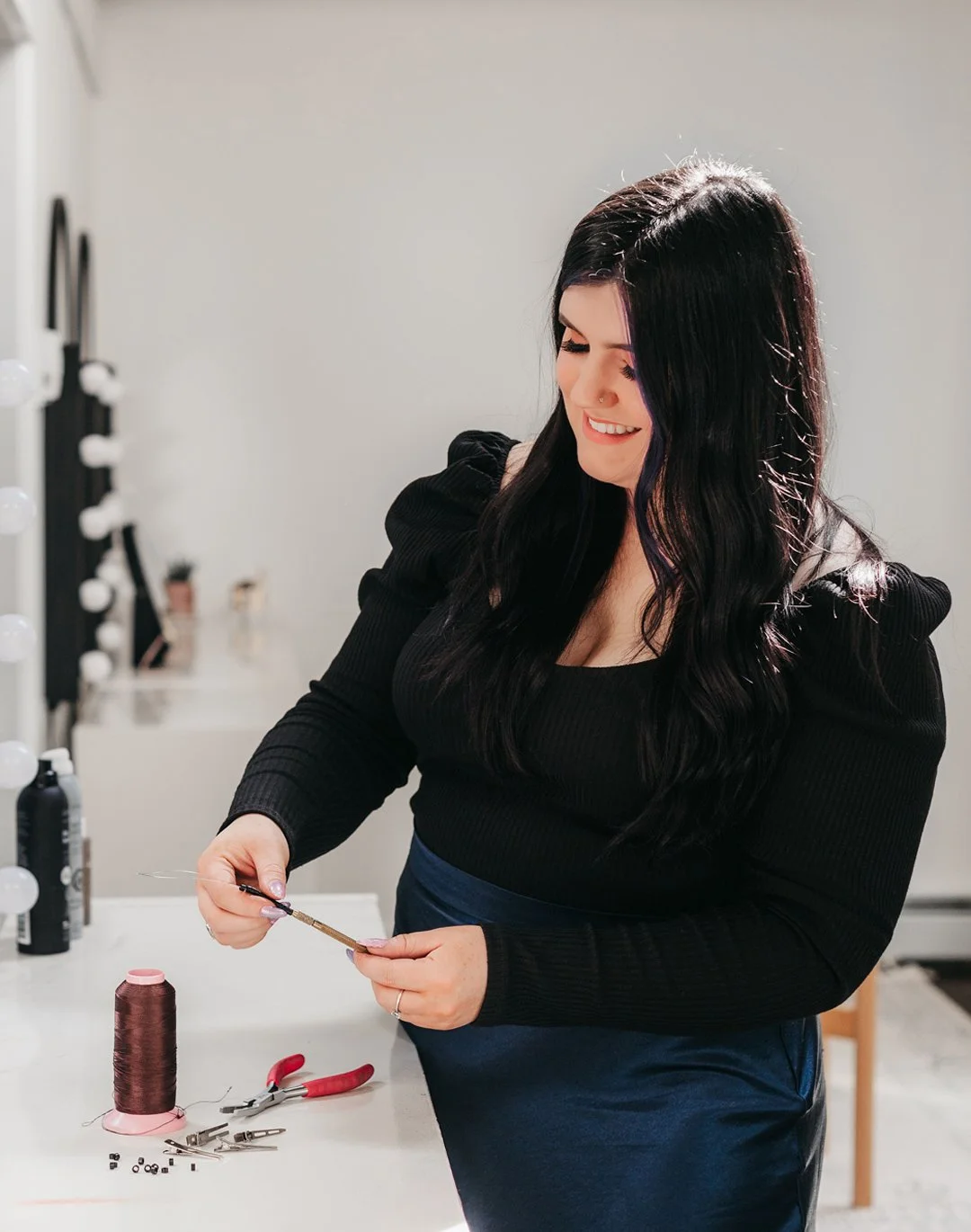 A woman with long dark hair, smiling, working with jewelry tools at a white table, with jewelry supplies including a spool of thread and pliers.