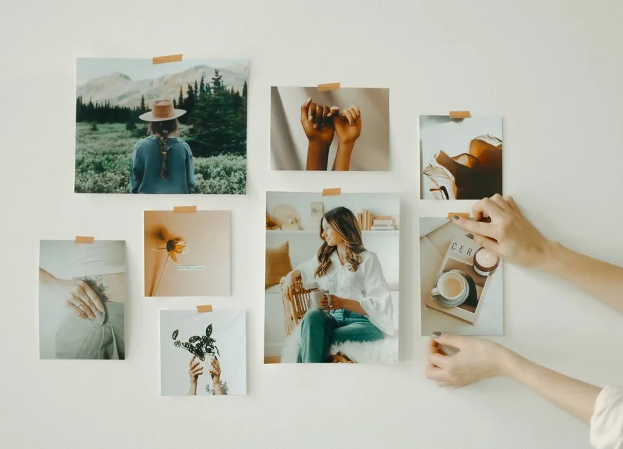 A collection of nine photographs taped to a white wall, including a woman in a hat in nature, a hand with a flower, two hands holding a small branch, a woman reading a book, a cup of coffee, and abstract images, with a person's hands arranging one of the photos.