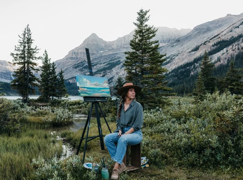 A woman sitting outdoors in a forested mountain landscape, with an easel and canvas displaying a partly cloudy sky scene, surrounded by trees and greenery.