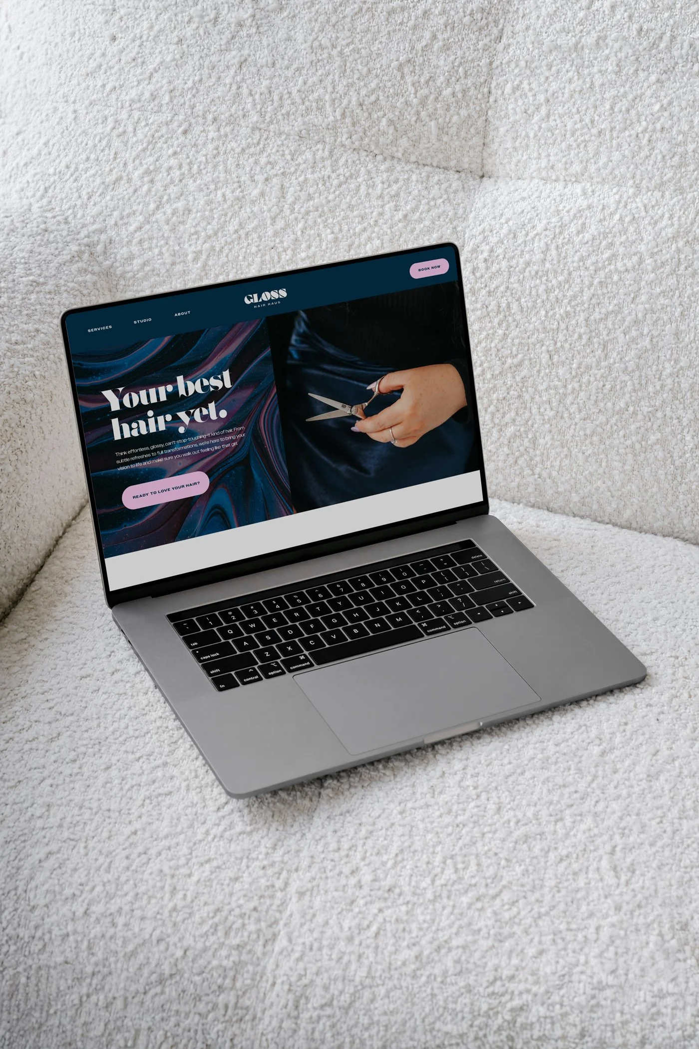 A laptop on a white textured surface, showing a website for a hair salon called 'GLOSS' with a slogan 'Your best hair yet' and a picture of someone holding scissors.