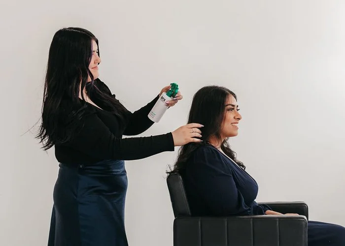 A woman is sitting in a salon chair while another woman sprays her hair with a styling product.
