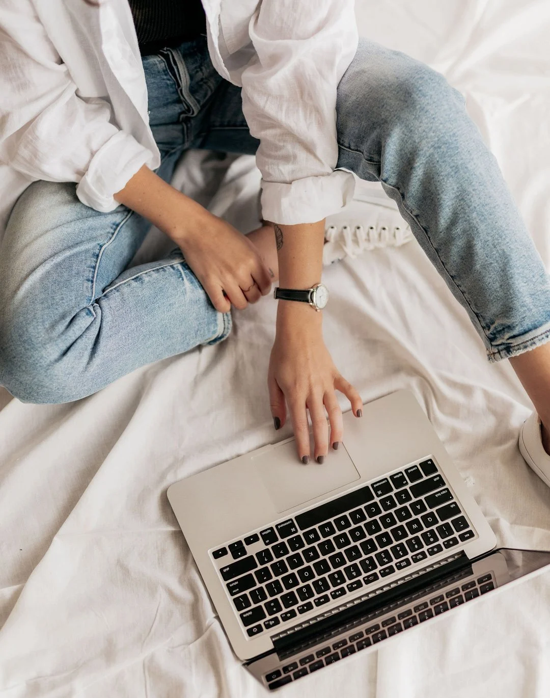 Person sitting on a bed with a laptop, wearing light blue jeans, a white long-sleeve shirt, and a watch, with their hand on the laptop's trackpad.