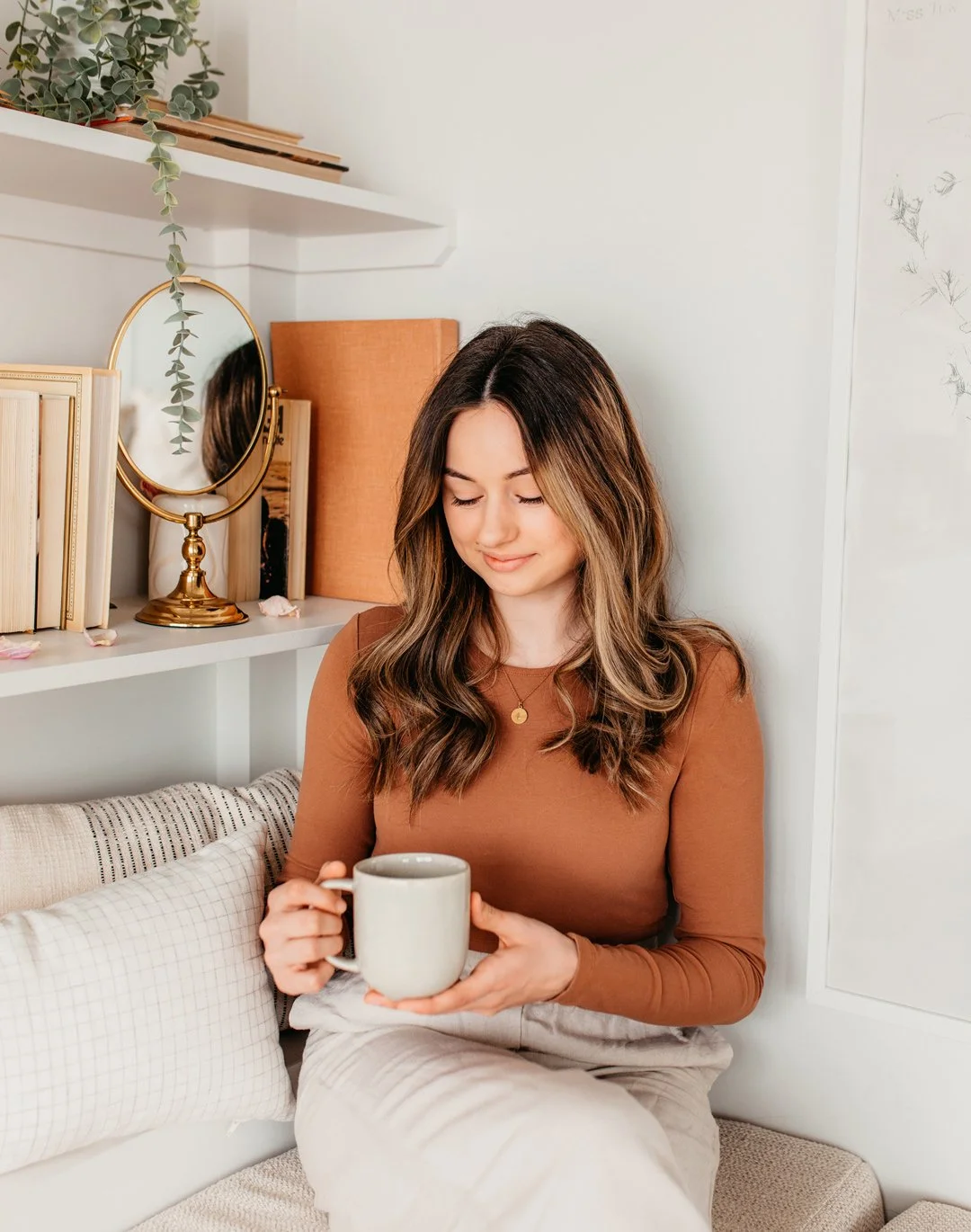 A woman sitting on a sofa holding a mug, with shelves, books, a mirror, and decorative items behind her.