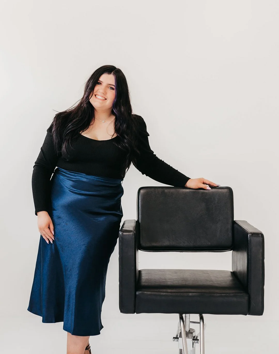 A woman with long dark hair, wearing a black top and a blue satin skirt, is smiling and leaning on a black salon chair in front of a plain white wall.