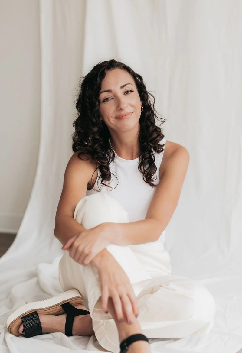 A woman with dark curly hair sitting on a white fabric backdrop, smiling with arms crossed, wearing a sleeveless white top and cream-colored pants, and black sandals.