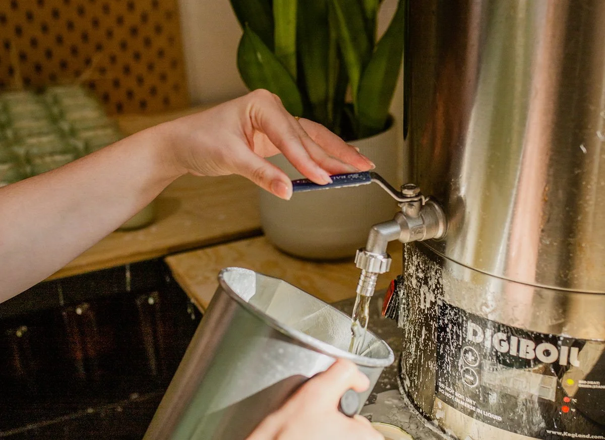 A person filling a stainless steel cup with hot wax from a hot wax dispenser. The person's hand is turning a blue lever.