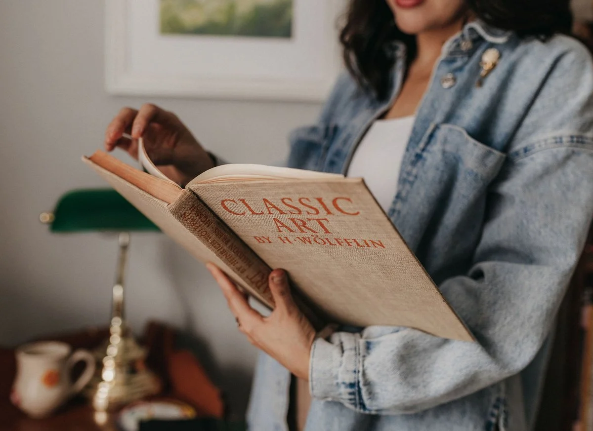 Person holding and reading a book titled 'Classic Art by H. Wolfflin' in an indoor setting with a green desk lamp and ceramic pitcher in the background.