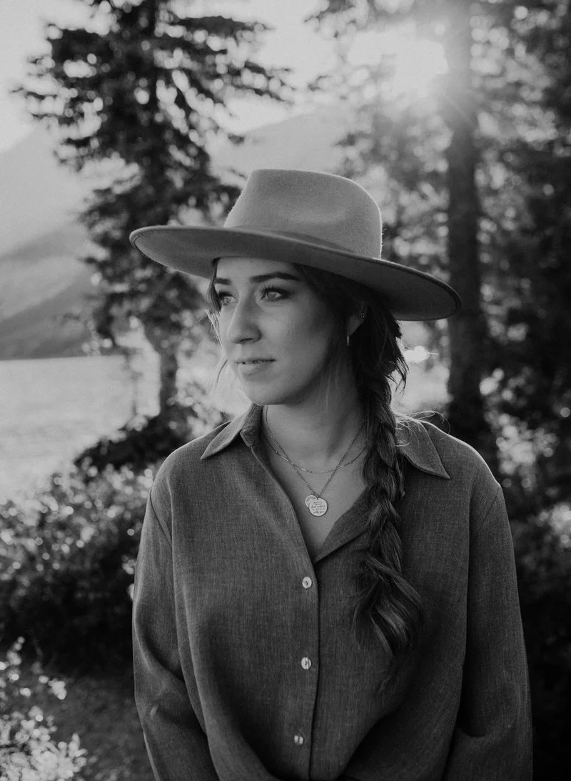 A woman wearing a wide-brimmed hat and a button-up shirt outdoors with trees and sunlight in the background.
