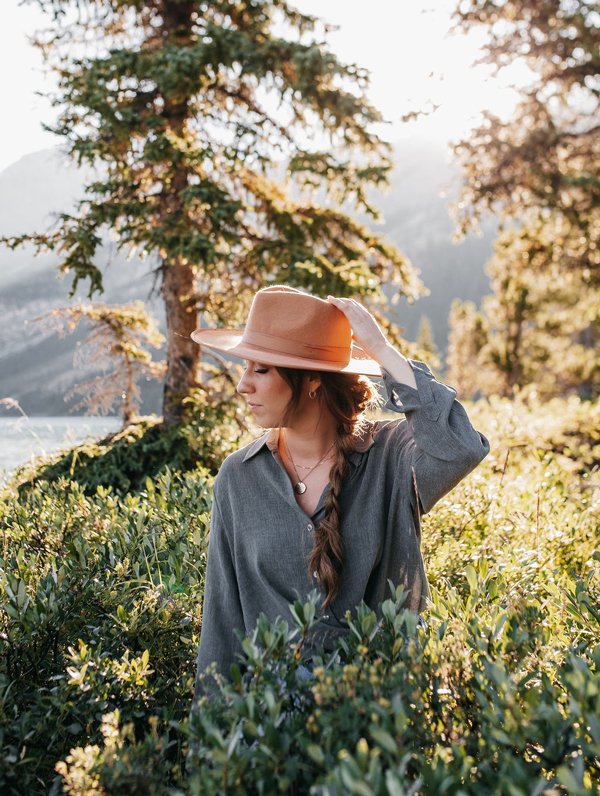 A woman with long hair in a braid, wearing a wide-brimmed hat, gray shirt, and necklace, standing in a sunlit forested area near water.