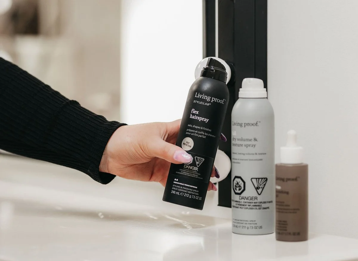 A hand holding a black bottle of Living Proof StyleLab flex hairspray with a gray cap, standing on a white bathroom countertop with two other Living Proof hair products in the background.