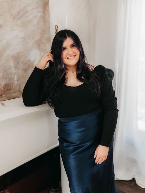 Young woman with long dark hair smiling, wearing a black top and a dark blue satin skirt, posing indoors by a white wall and curtains.