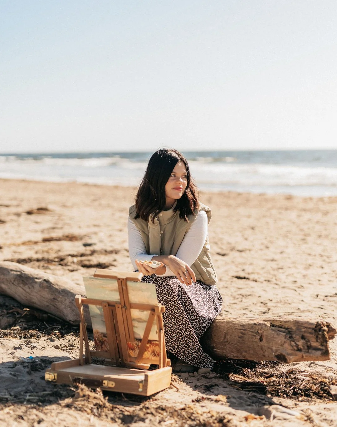 A woman sitting on a beach next to a large log, with an open wooden artist's easel in front of her, looking softly into the distance, with the ocean and sky in the background.