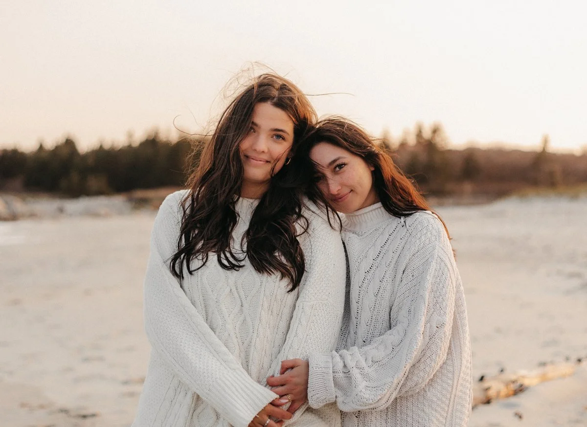 Two women in white sweaters hugging on a beach at sunset.