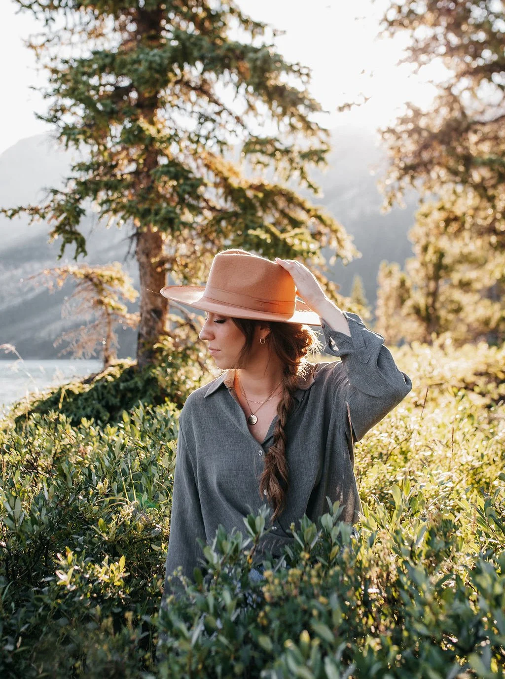 A woman in a gray shirt and a tan wide-brimmed hat stands among green bushes in a scenic outdoor setting with trees and mountains in the background, illuminated by warm sunlight.