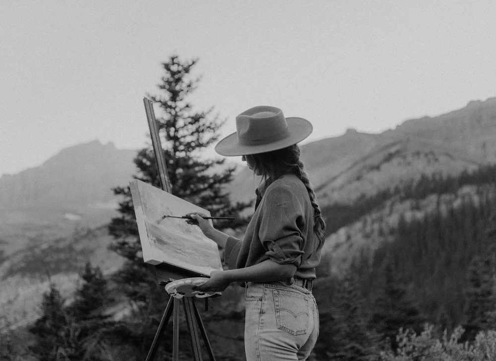 A woman painting outdoors with mountains and trees in the background, wearing a hat and casual clothes.