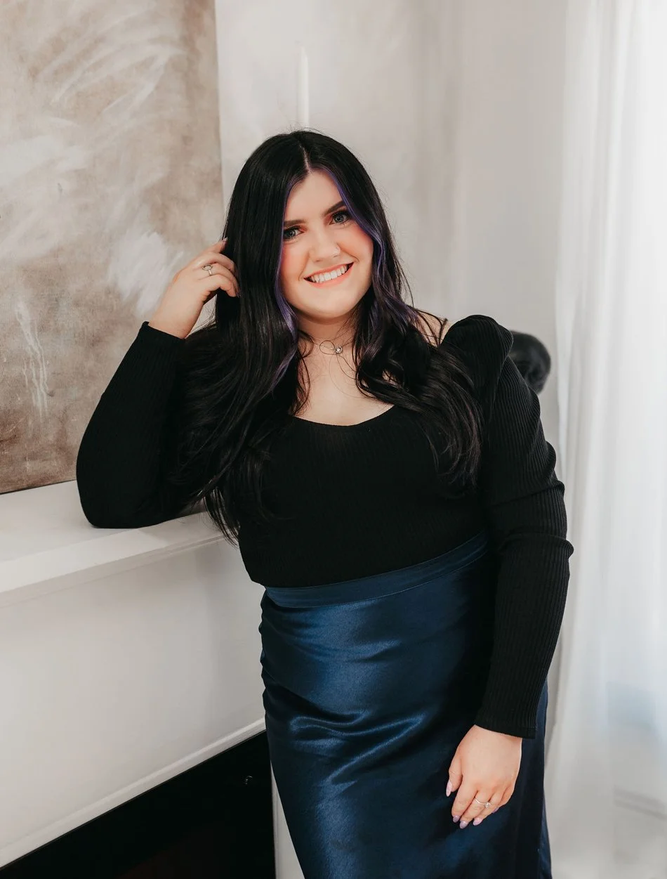 A woman with long black hair, some purple streaks, and a smile, posing indoors near a white wall and curtain, dressed in a black top and a dark blue satin skirt.