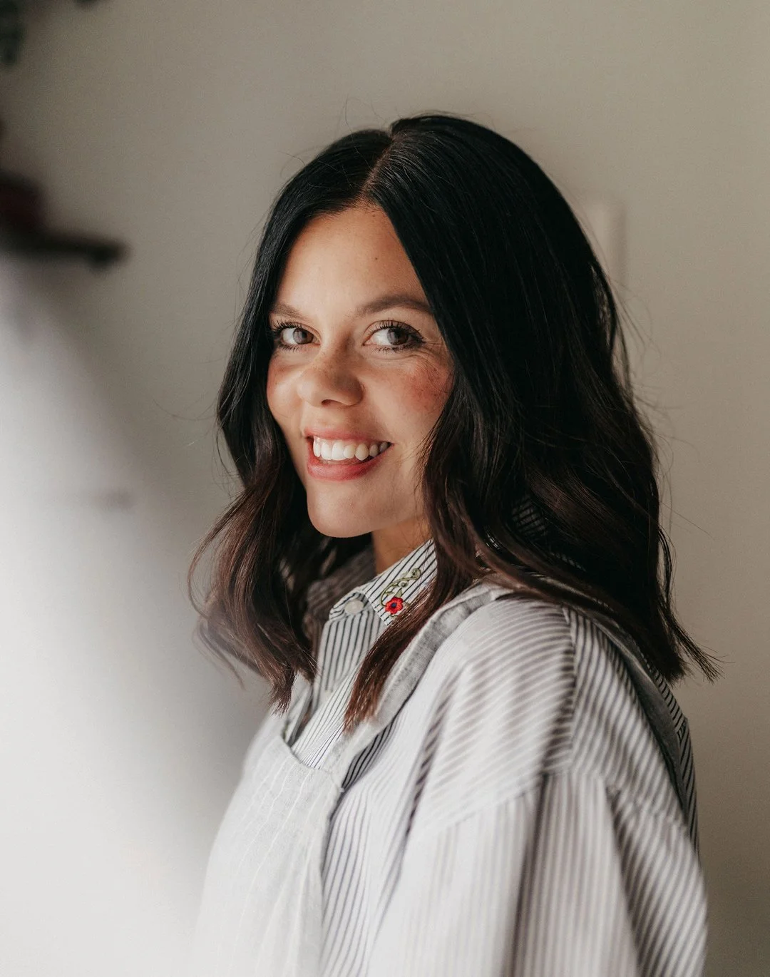 A woman with shoulder-length dark hair smiling at camera, wearing a light striped shirt with embroidered flowers near the collar, indoors with a neutral background.