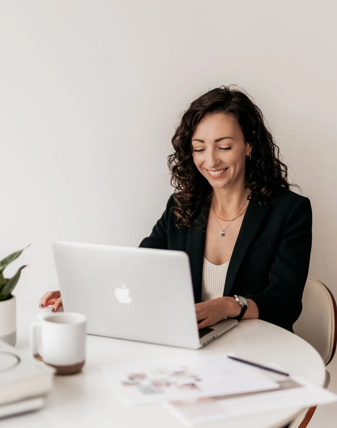 A woman with curly dark hair sitting at a white desk, smiling while using a silver MacBook, with a white mug, some papers, and a small potted plant nearby.