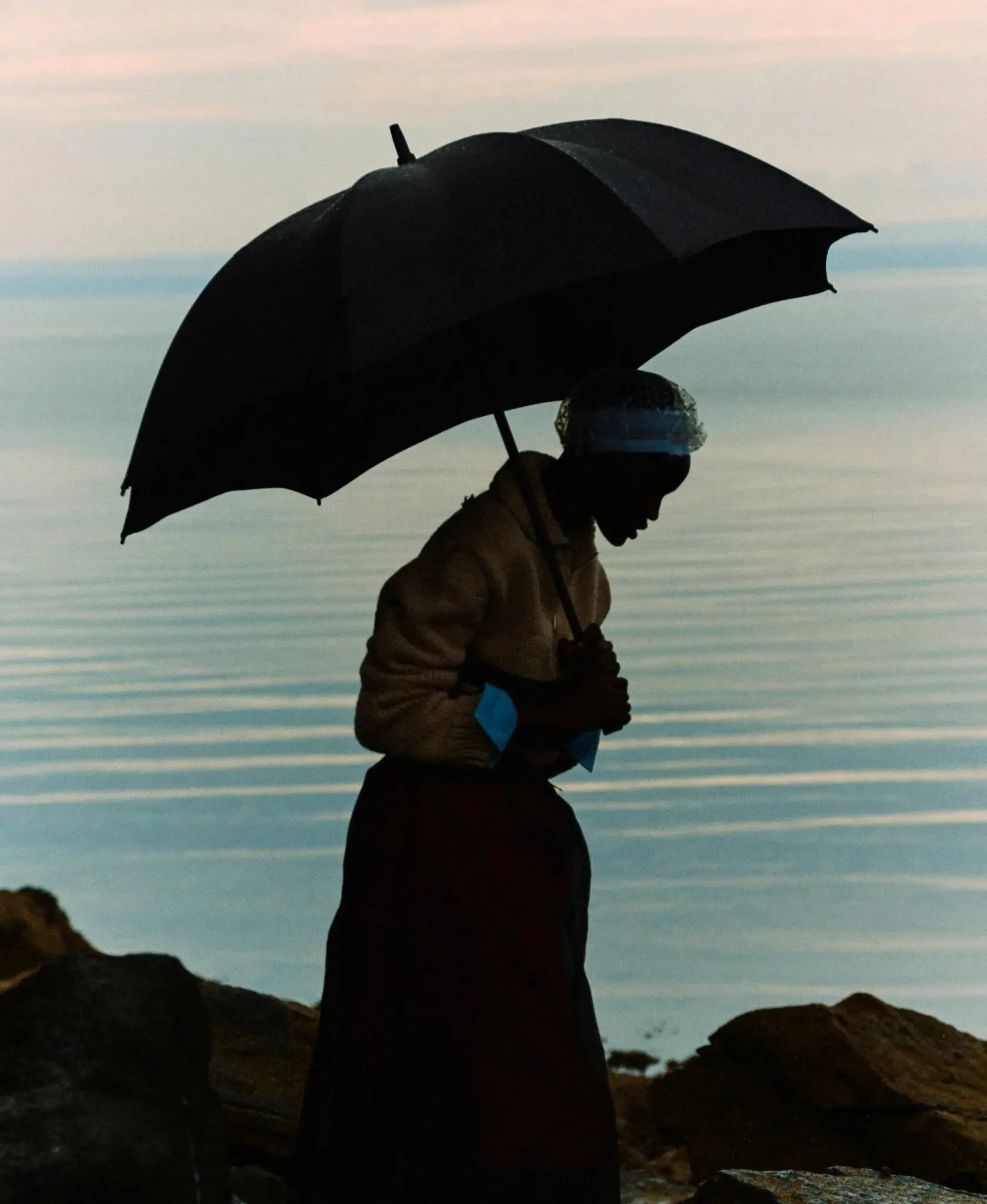 Female model with umbrella silhouetted in front of a serene Scottish coastline. Photographed in Scotland by Jack Davison for T: The  New York Times Style Magazine. 

Model wears Prada sweater, shirt, skirt and skirt (worn underneath); and stylist’s o