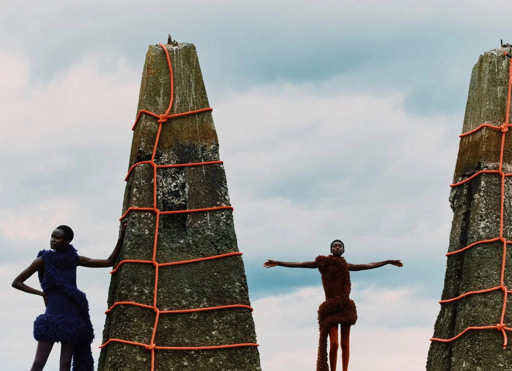 Two models pose next to cylindrical concrete structures with red rope and cloudy sky.  
Clothing from left: Loewe dress and stylist’s own tights. Loewe dress; and stylist’s own tights.

Credit...Photograph by Jack Davison.
Production by Mini Title 
L