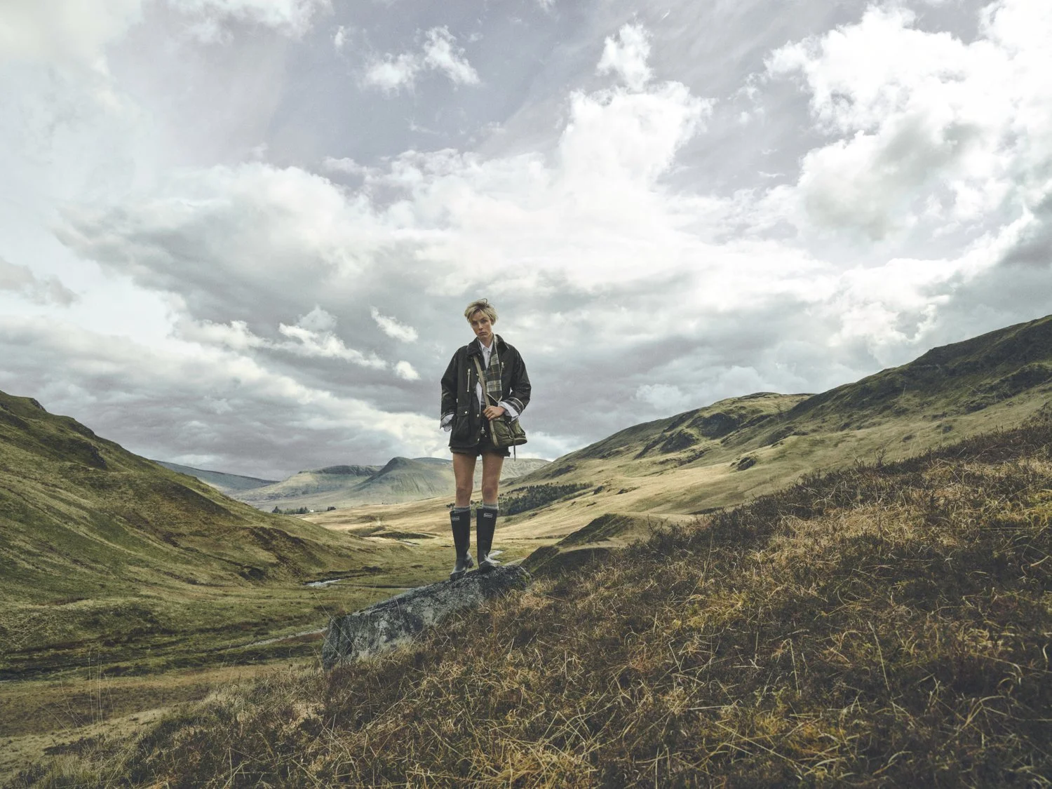 Model, Edie Campbell, stands on a rocky outcrop in the Scottish Highlands with mountains behind her. Shot for Barbour Spring Summer 2026 campaign by Dan Jackson