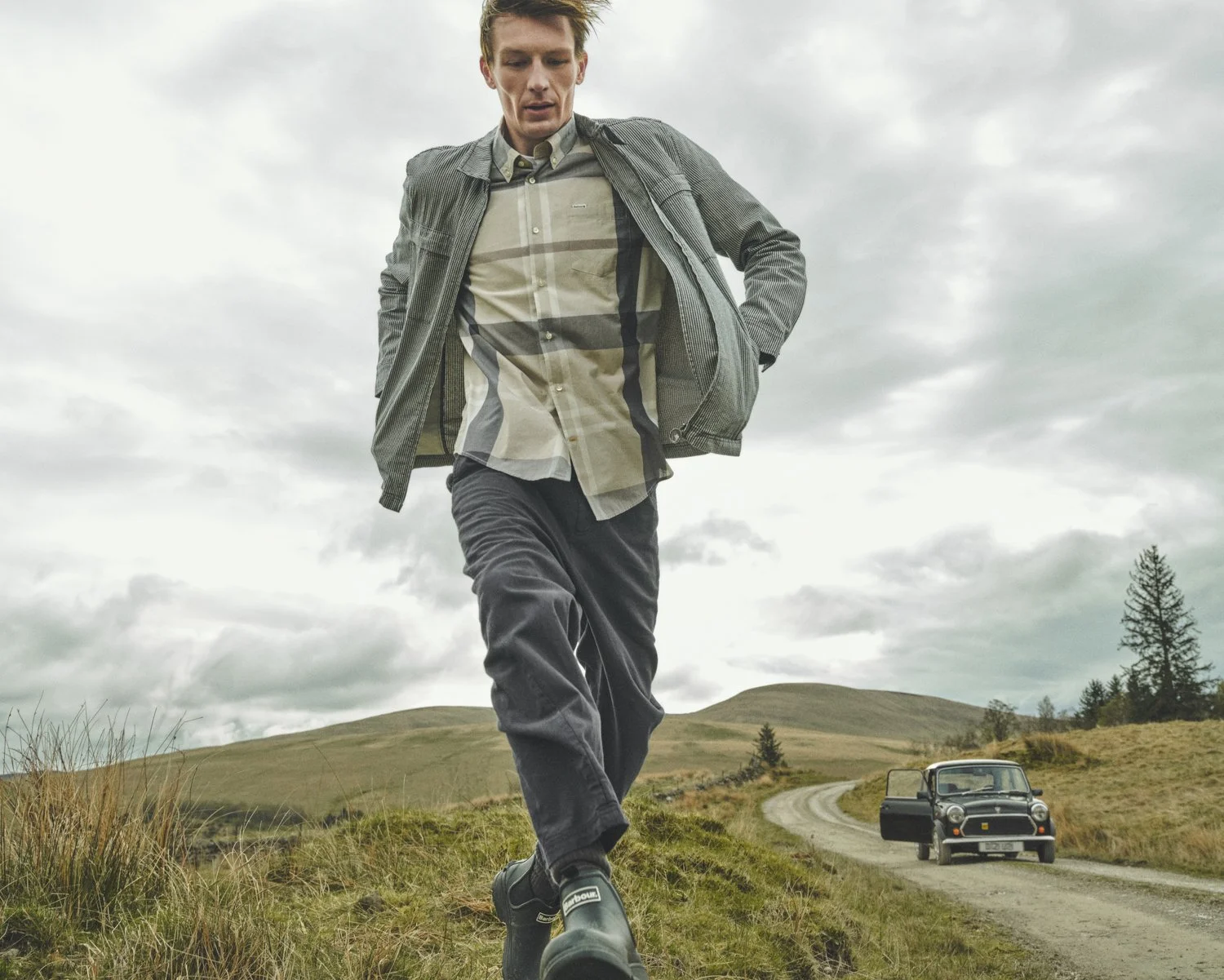 Finnlay Davis walks with a classic mini cooper as a prop behind him whilst modelling for  for Barbour SS26 in the  Scottish Highlands