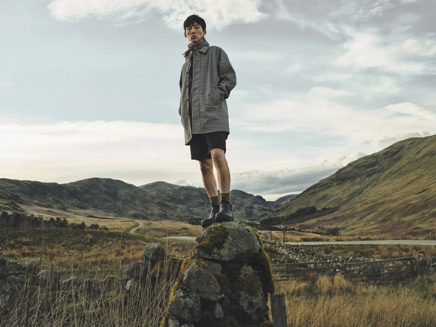 Dan Jackson photographs Woo Sang for Barbour SS26 who is standing on an old stone wall with mountains and Scottish countryside behind