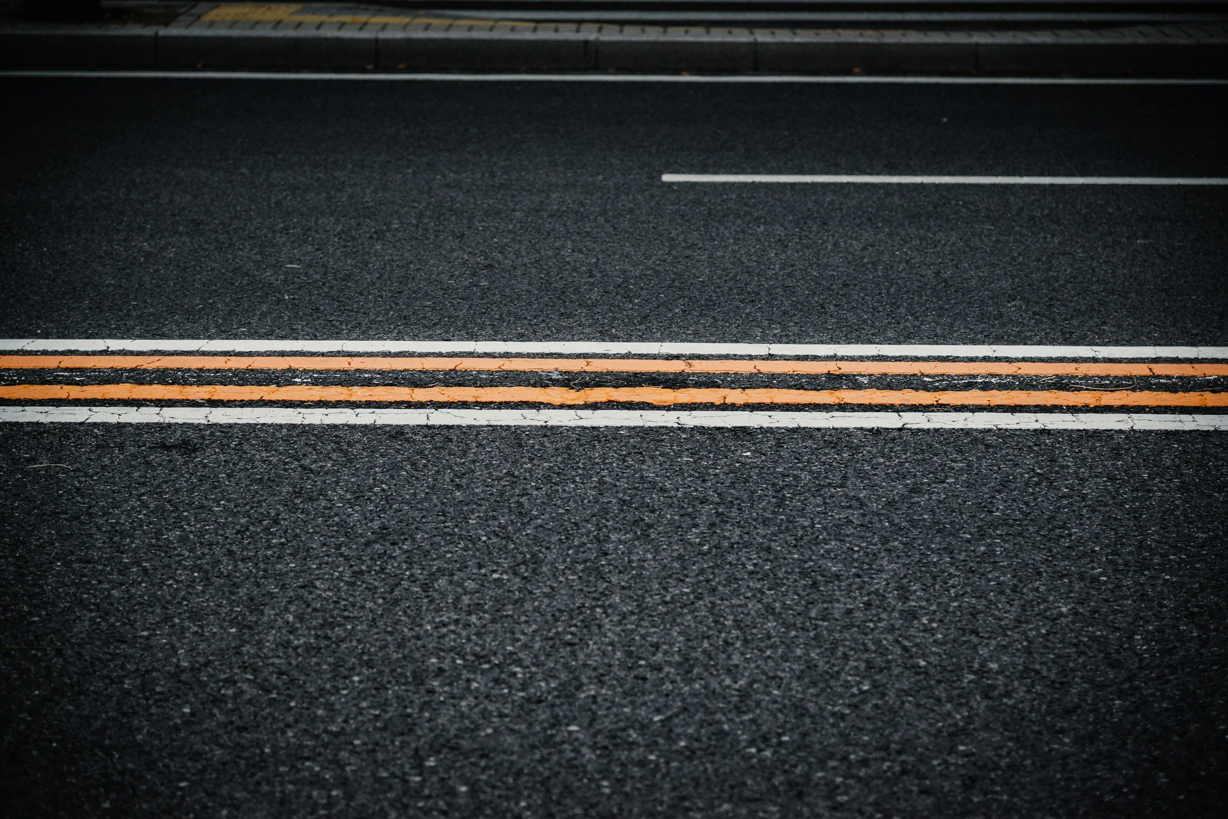 Close-up of a black asphalt road with painted white, orange, and yellow lines at the bottom of the image, with a curb and sidewalk in the background.