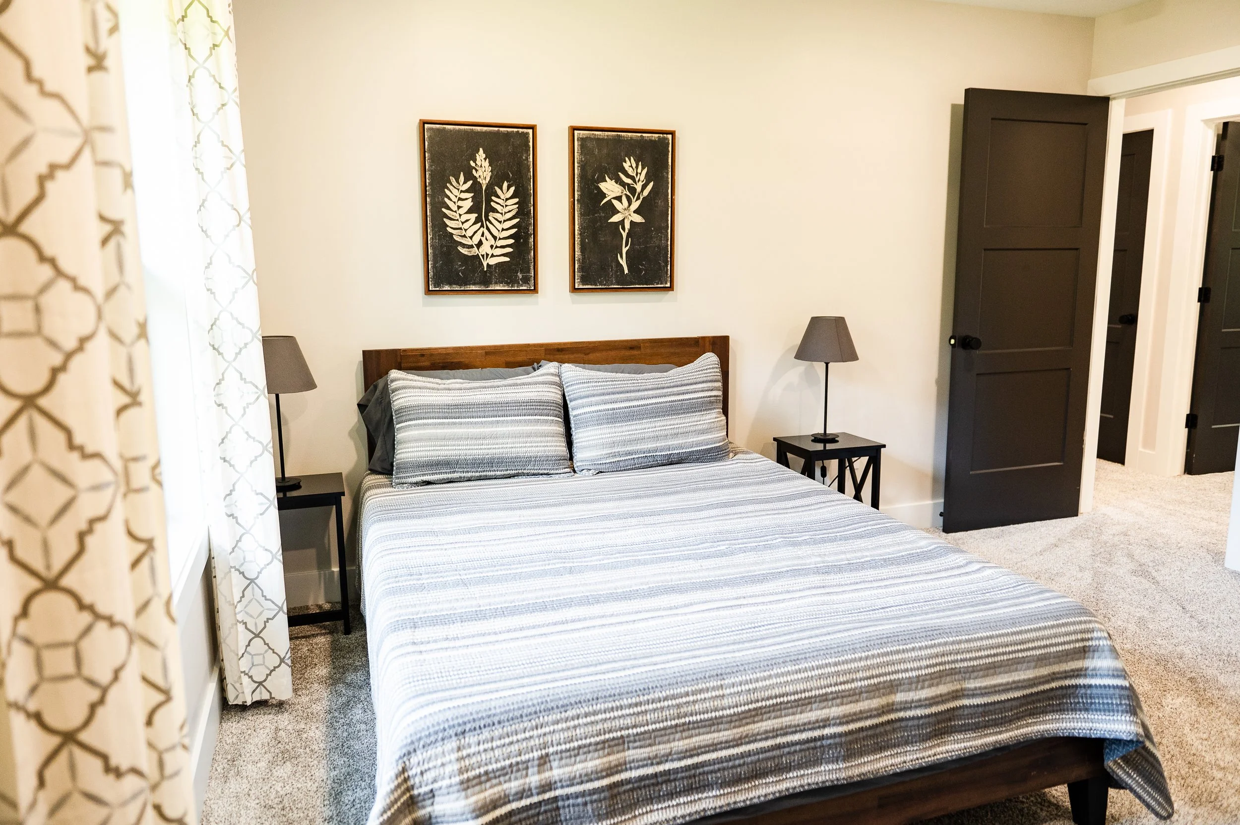 A bedroom with a bed covered in striped gray and white bedding, flanked by two black nightstands each with a gray lamp. Two framed botanical prints hang above the headboard, and a black door is visible on the right side. A patterned curtain partially