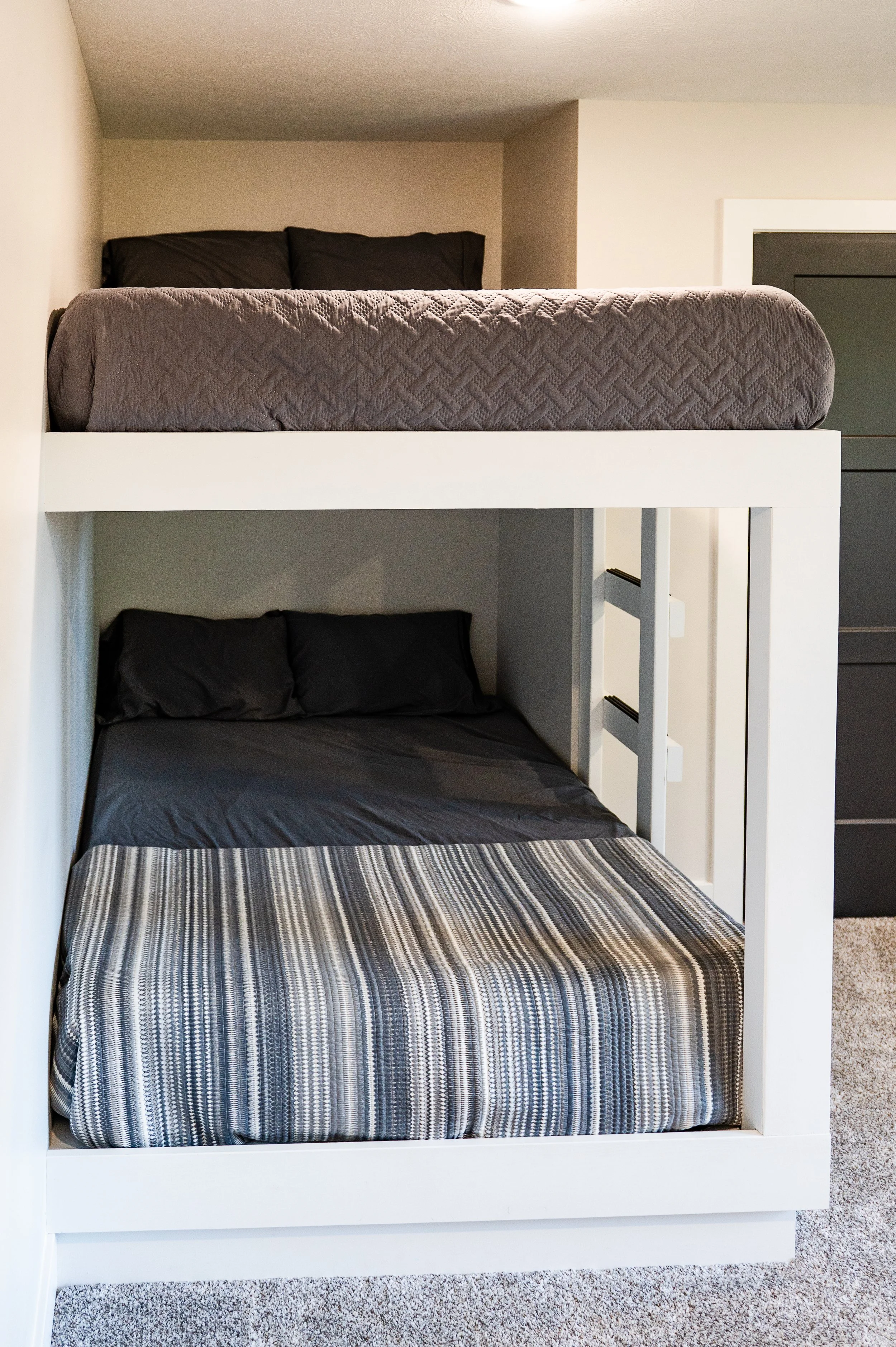 A white bunk bed with a twin bed on top and a full bed on the bottom in a bedroom. Both beds have dark gray bedding and pillows. The top bed has a textured quilt, and the bottom bed has a striped bedspread. The room has beige walls and a carpeted flo