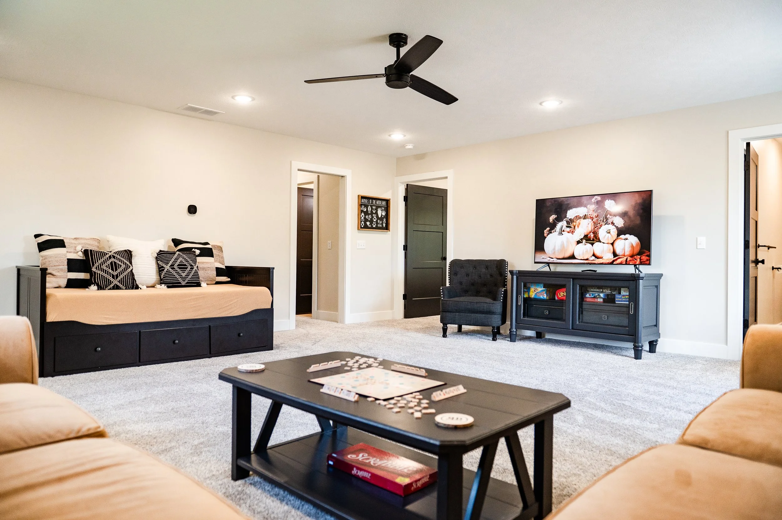Living room with beige walls and carpet, featuring a large black and beige sofa with patterned pillows, a black armchair, a dark gray media console with a large pumpkin-themed picture on the wall above, a black coffee table with board games and a rem