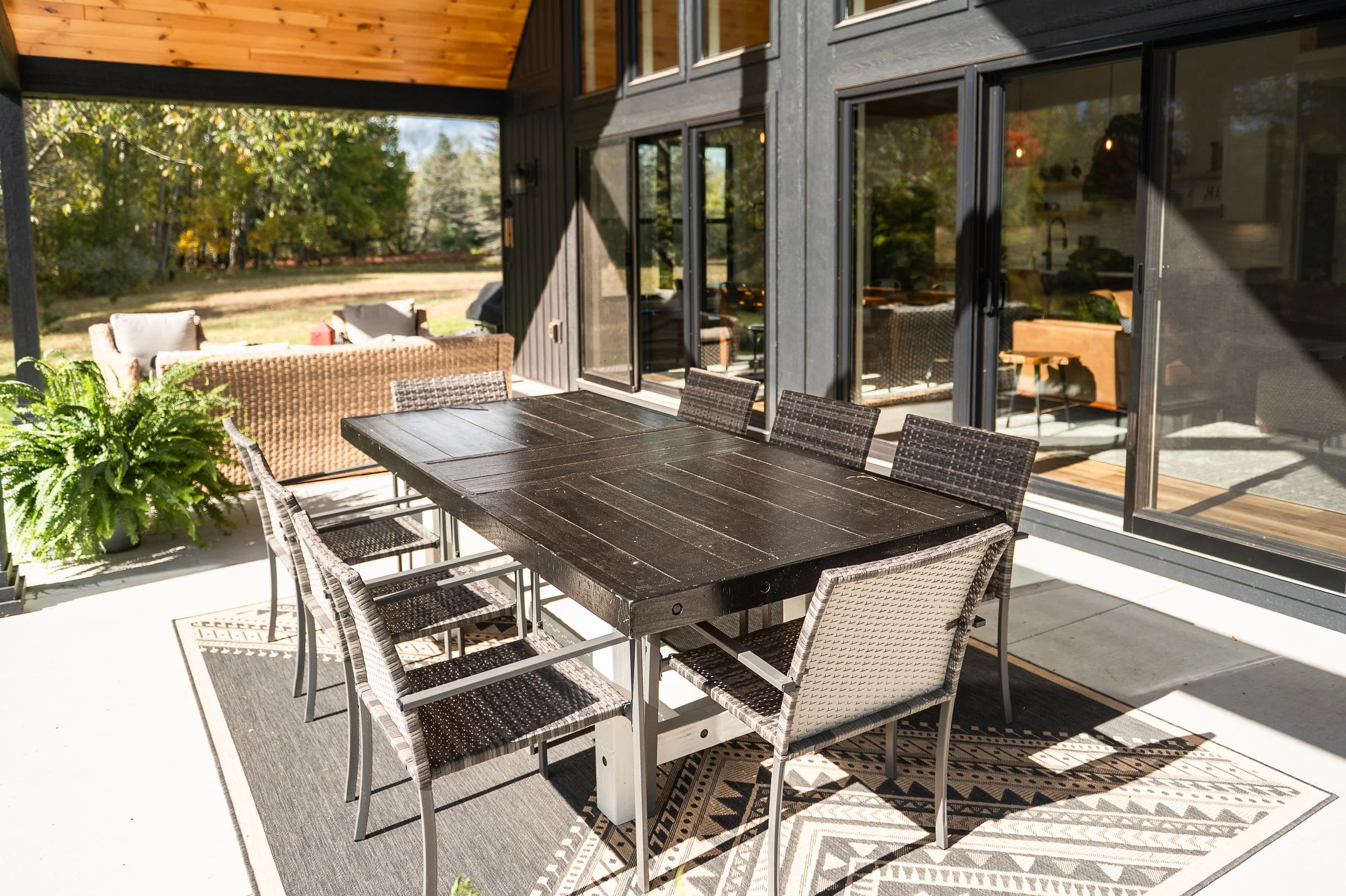An outdoor patio area with a black dining table and eight matching chairs, a rattan sofa with cushions, and greenery in the background. The patio is shaded with sunlight, and the house has large glass sliding doors.