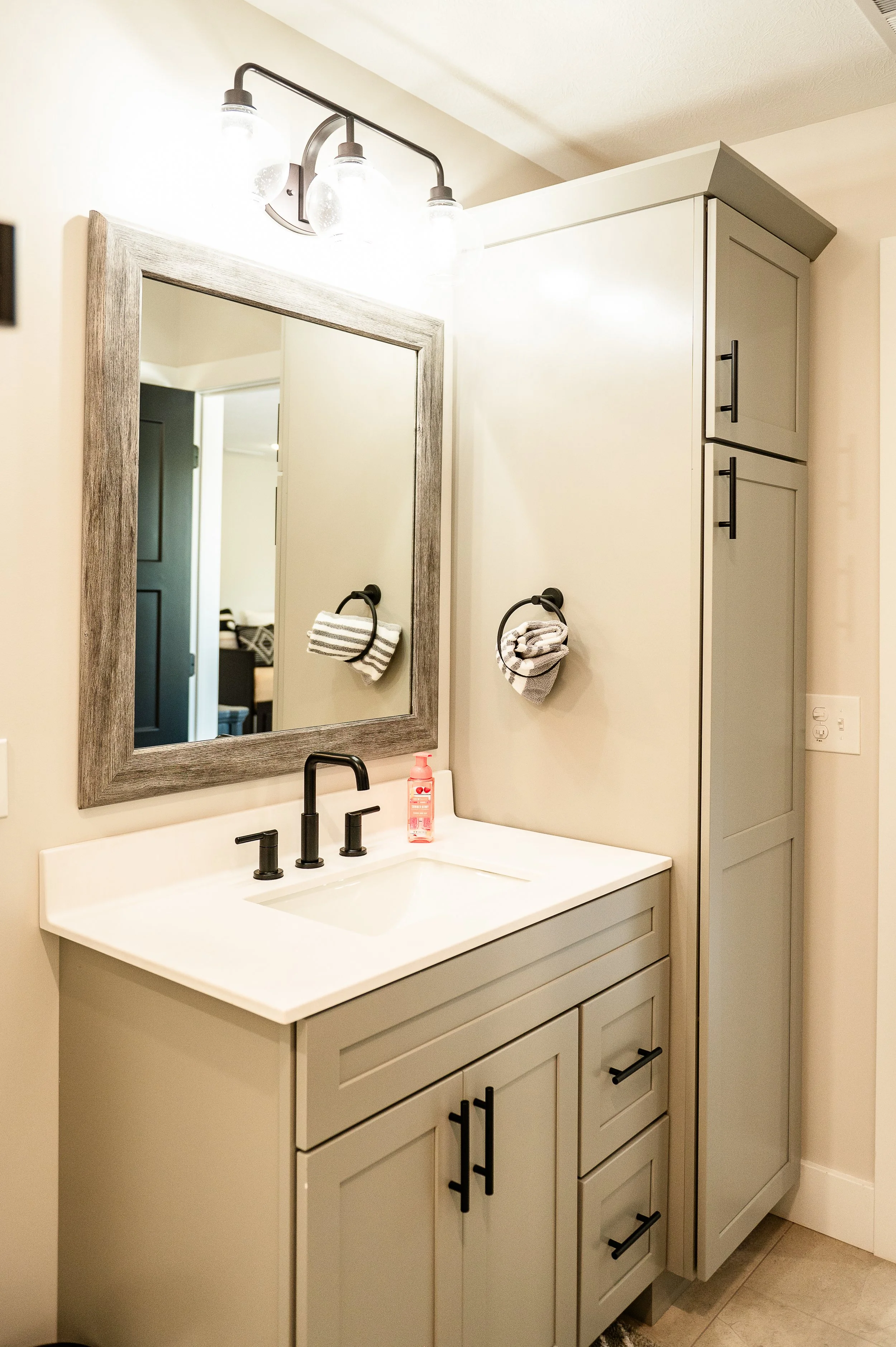 Bathroom vanity with a white countertop, black faucet, mirror with a gray frame, gray cabinets, and black hardware. Two striped hand towels hang on black towel rings, with a pink soap dispenser on the counter.