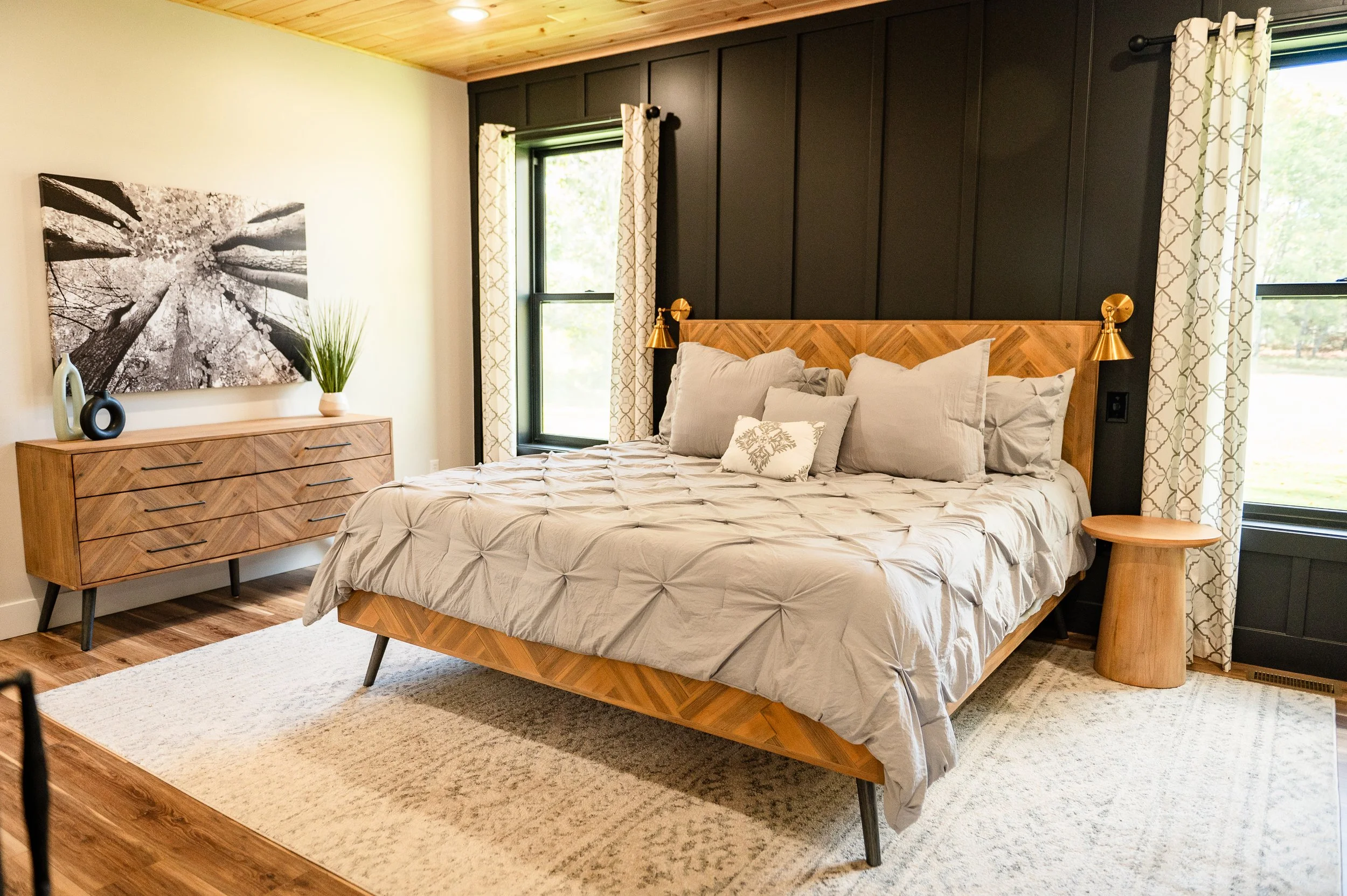 A bedroom featuring a bed with a wooden headboard and multiple pillows, two gold wall-mounted lamps, a wooden dresser with decorative vases and a plant, framed artwork on the wall, dark window frames with patterned curtains, and a beige rug on wood f