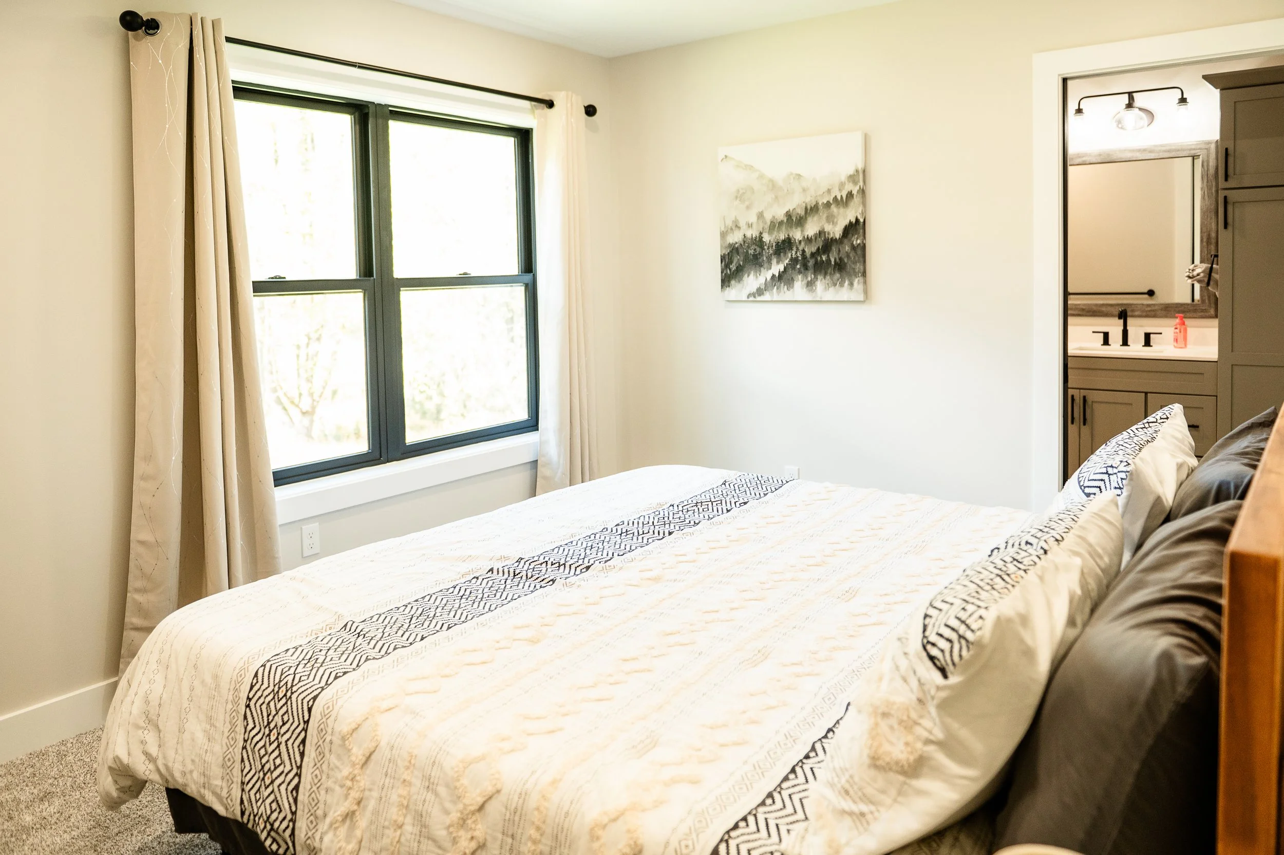 A bedroom with a bed covered in a cream-colored and black patterned quilt, dark pillows, a large window with beige curtains, and a framed black-and-white mountain landscape painting on the wall. There's a doorway to a bathroom with a gray vanity, mir