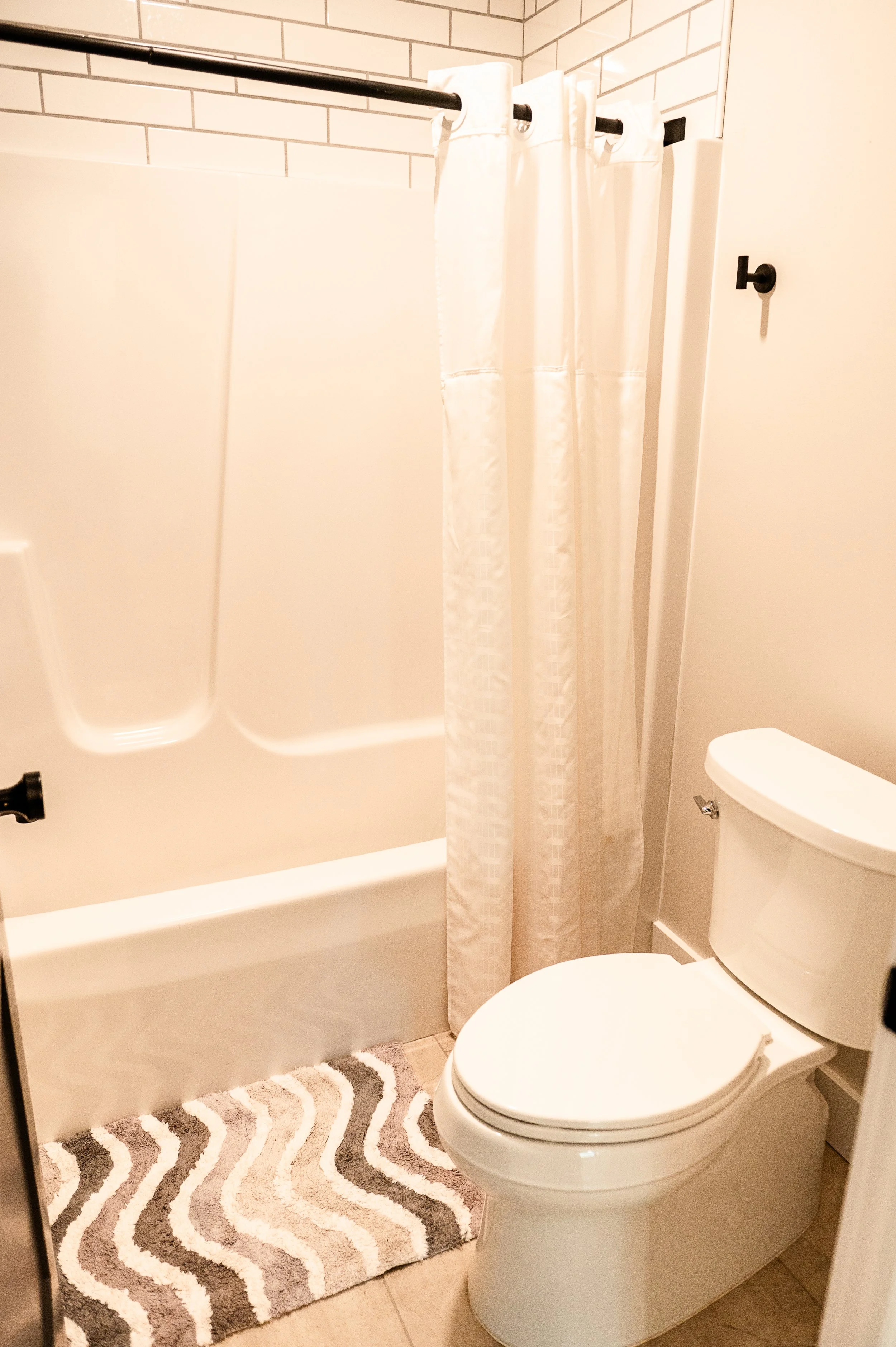 A bathroom with a white bathtub and shower area featuring a shower curtain, a toilet, and a rug on the beige tiled floor.