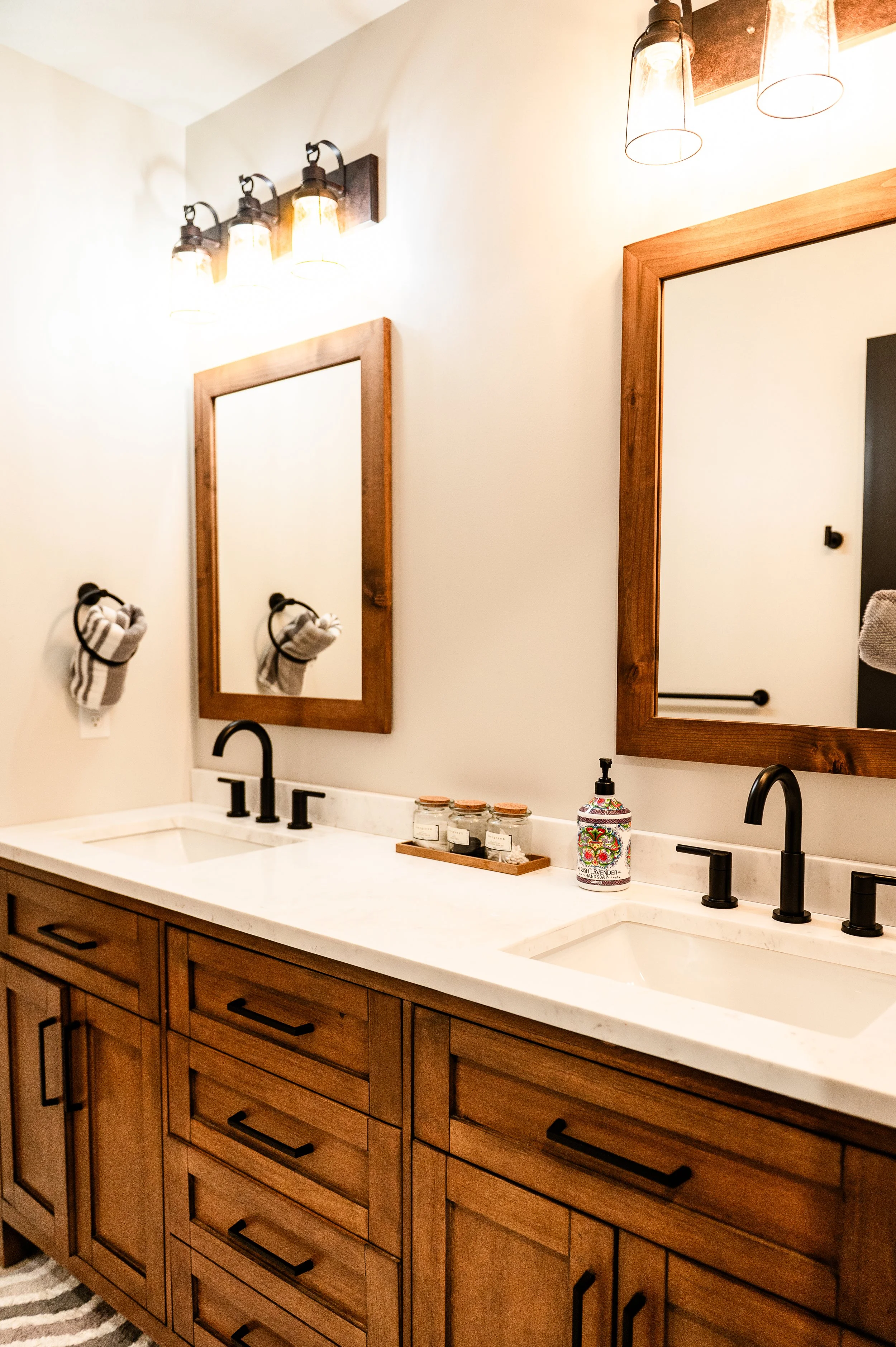 A bathroom vanity with a white countertop, two sinks, black faucets, and wooden cabinets. Above the vanity are two large rectangular mirrors with wooden frames. To the left, two black towel rings hold folded towels, and on the countertop are containe