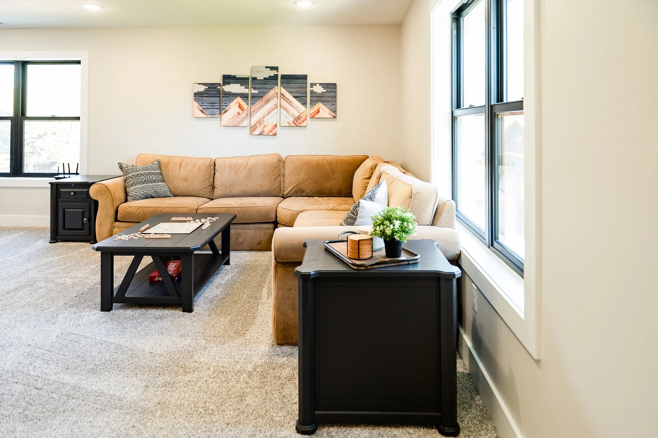 Living room with beige couch, black side tables, a coffee table with a Scrabble game, and large windows. Wall art of mountain peaks is above the couch. Potted plant on side table.