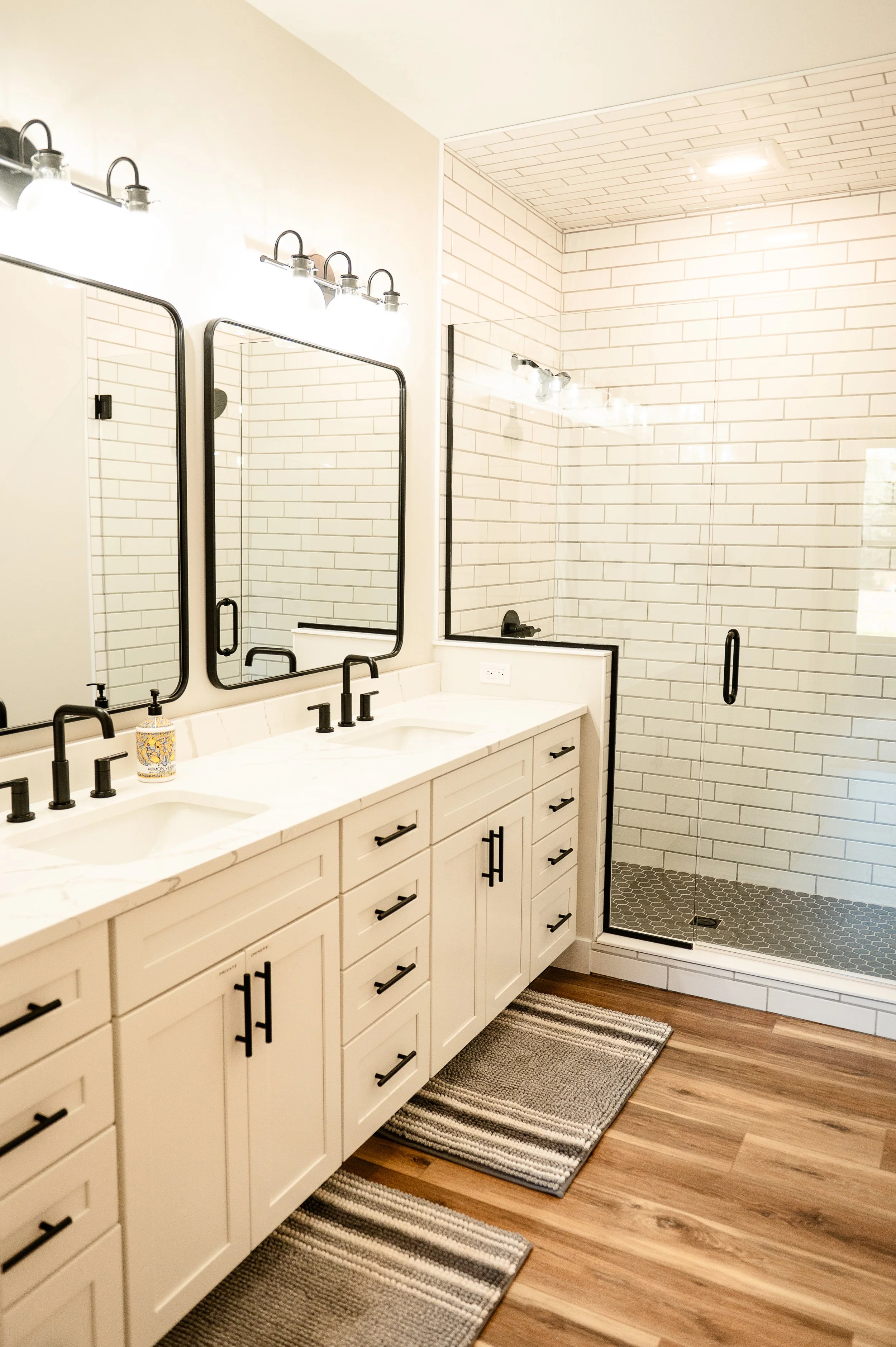 Modern bathroom with a double vanity with white cabinets and black handles, a large wall mirror with black frame, black fixtures, and a glass shower with white subway tile walls and a black framed door. Wood flooring and striped rugs.