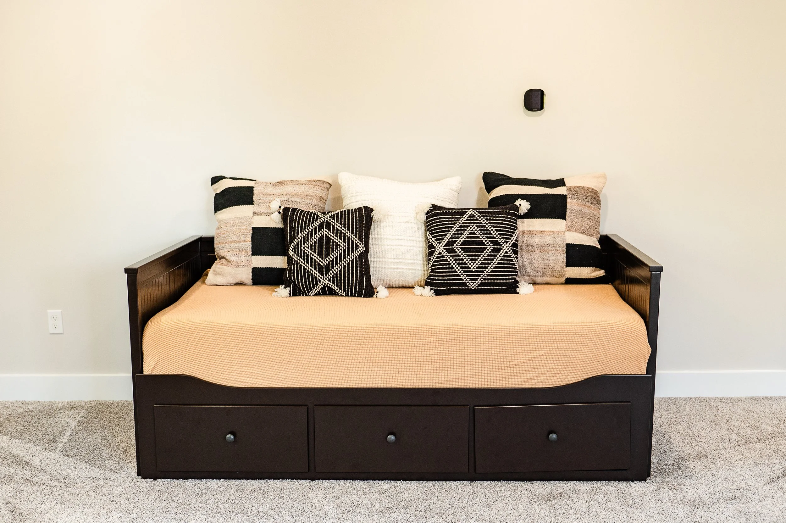 A black bed frame with a drawer at the bottom, a beige bedspread, and five decorative pillows with black, white, and beige geometric patterns, against a plain off-white wall with a black wall-mounted device.