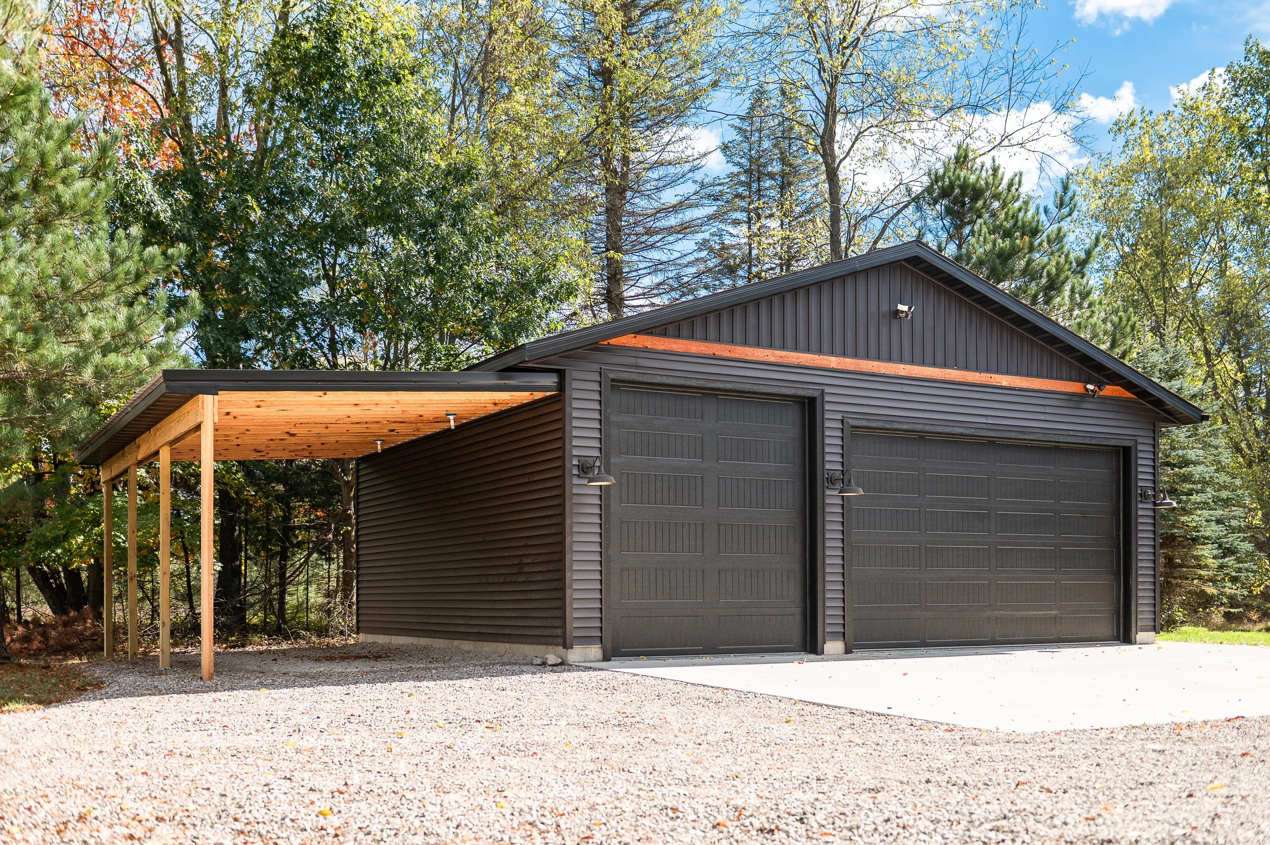 A two-car garage with dark gray siding and a red shingle roof, surrounded by tall trees with green and yellow leaves, under a bright blue sky with a few clouds.