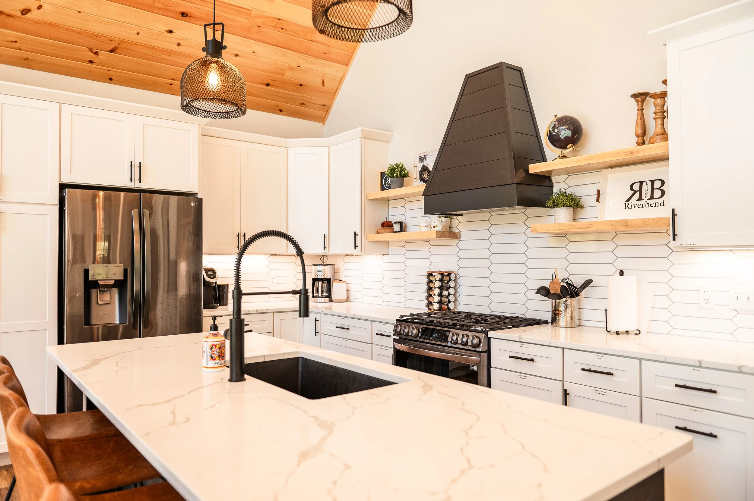 Kitchen with white cabinets, a stainless steel refrigerator, a black vent hood over a stove, a wooden shelf with decorative items, and a marble island with a black faucet.