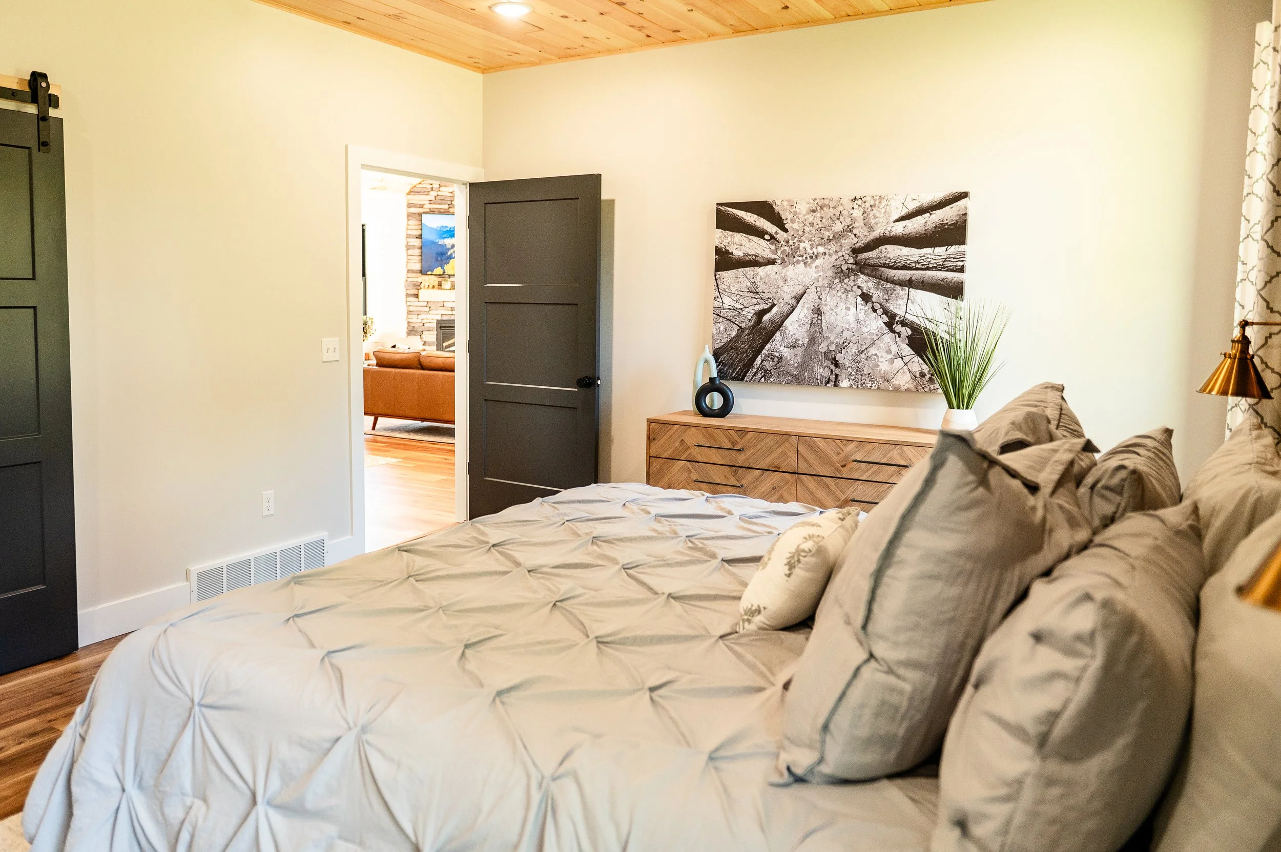 A bedroom with a bed covered in a beige quilt and pillows, a dark wood dresser, a large black and white forest-themed wall art, and a small plant in a white vase. An open door leads to a living room with a brick fireplace and a painting of mountains.