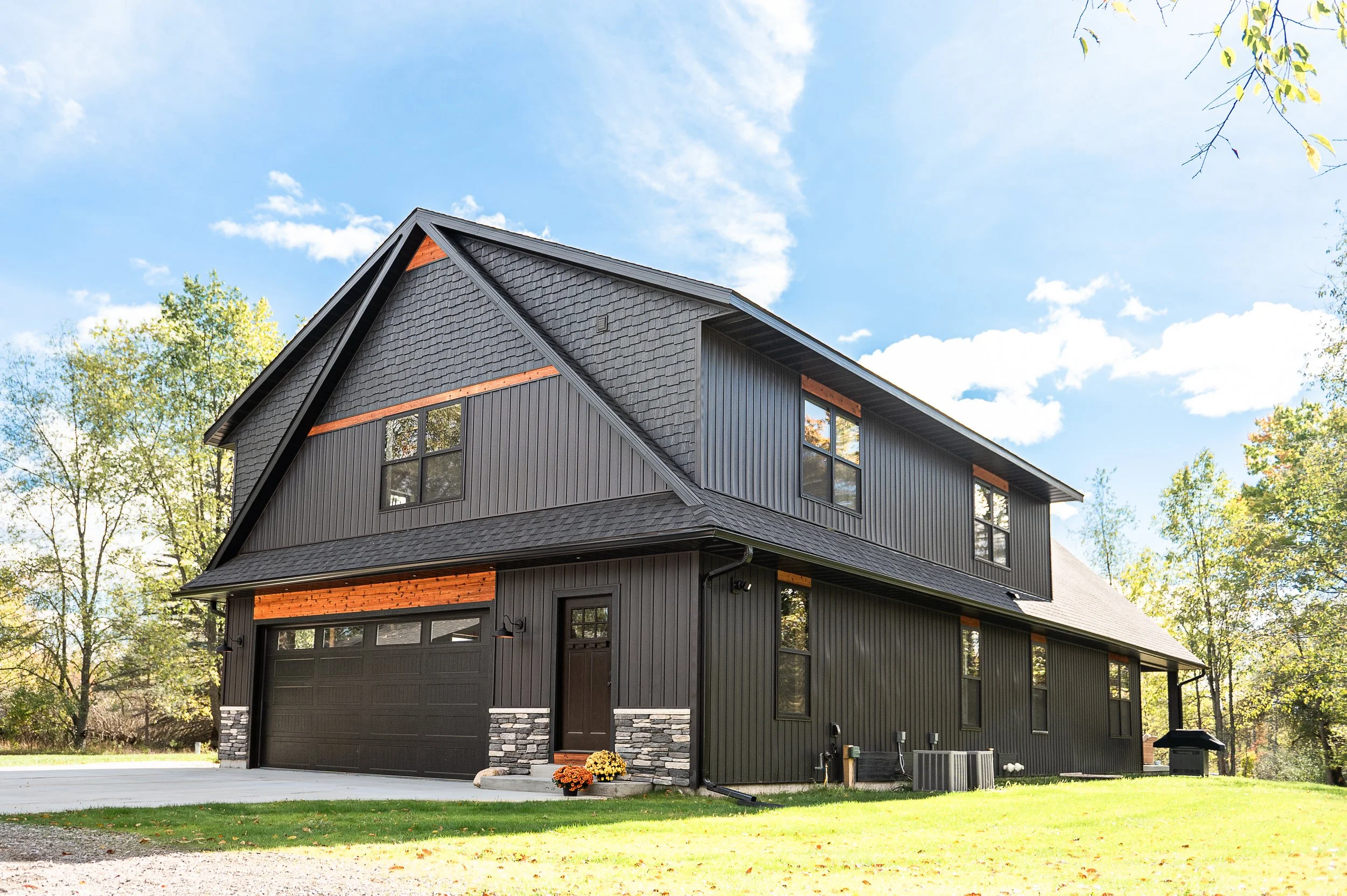 A modern house with black siding and a steeply pitched roof, surrounded by trees with fall foliage, under a blue sky with clouds.