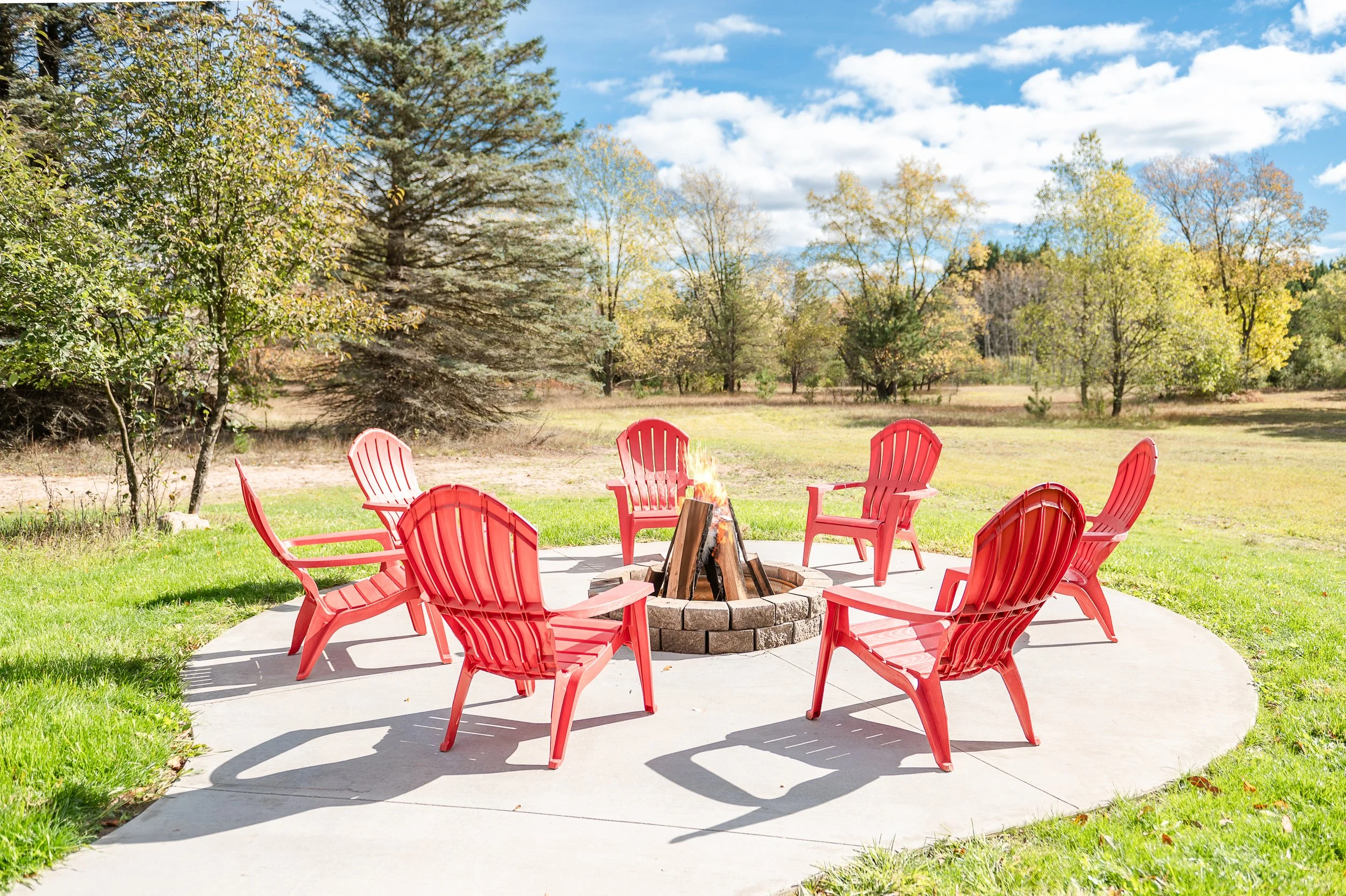 Red outdoor chairs arranged in a circle around a fire pit on a concrete patio, set against a backdrop of trees and a partly cloudy sky.